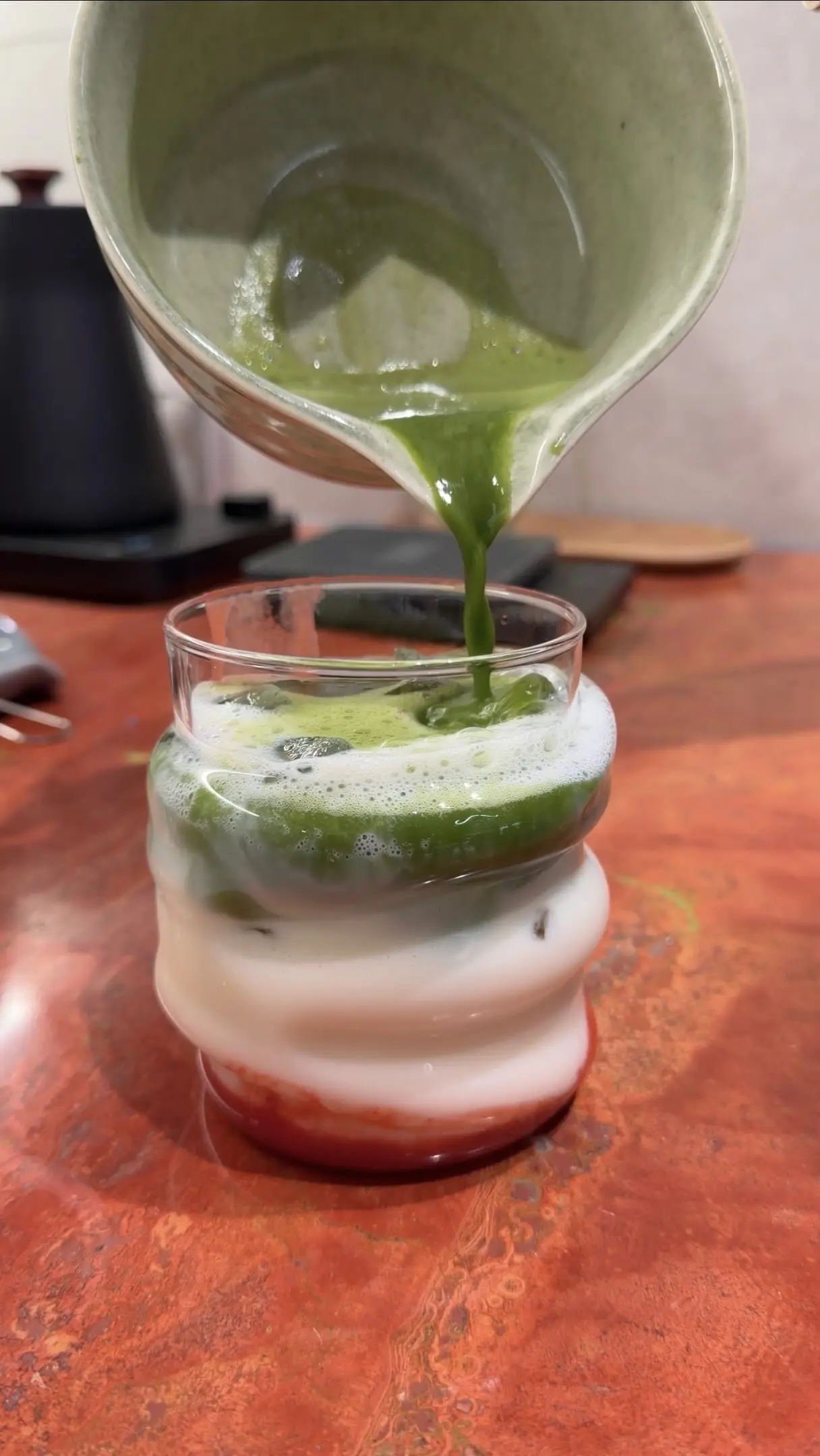 Matcha wird aus einer Glaskanne über Milch und Erdbeerpüree gegossen